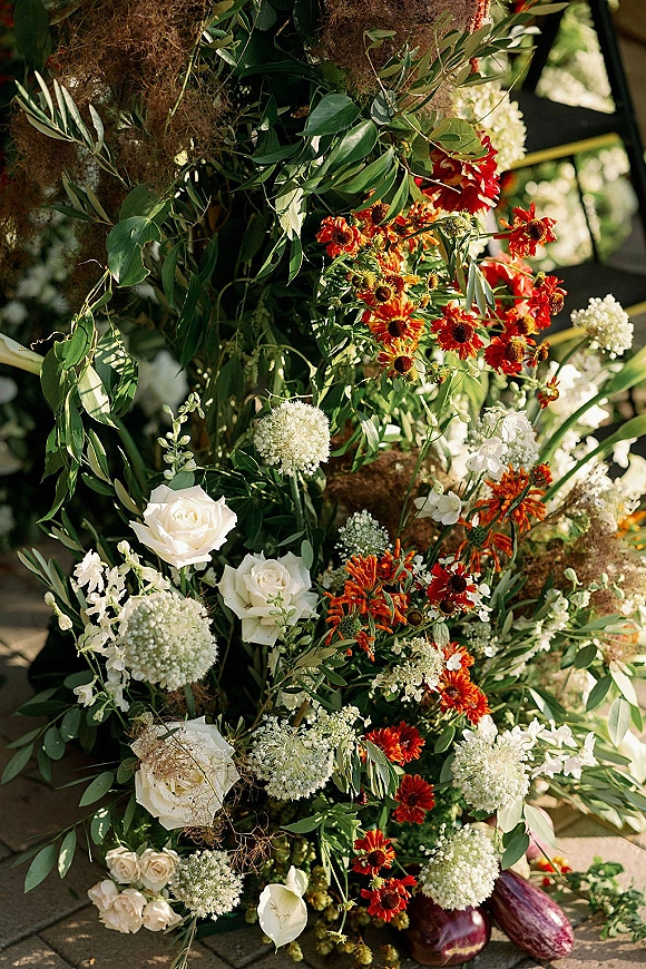 Wedding floral arrangement with ceremony flower installation styling, white roses and orange daisies with greenery and dried grasses on sunlit pavement