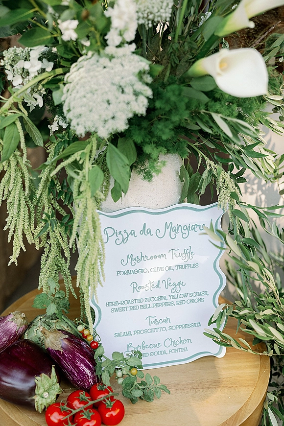 Wedding menu sign with calla lilies and greenery in a vase, scalloped menu card beside eggplants, tomatoes, and herbs on a wood table