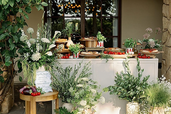 Wedding food station featuring a wedding pizza station on a white buffet table with greenery garlands, white florals, and menu sign on an outdoor patio