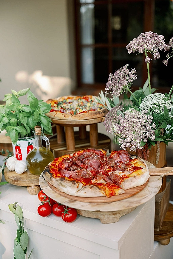 Wedding food table with pizza on wooden serving boards, basil and garlic accents, cherry tomatoes, olive oil, and florals by a window