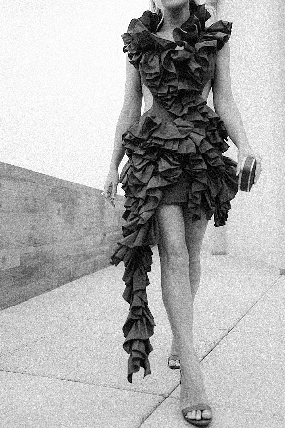 Reception dress in a black wedding reception dress style with ruffle mini hem, clutch purse and high heel sandals on a concrete walkway under open sky