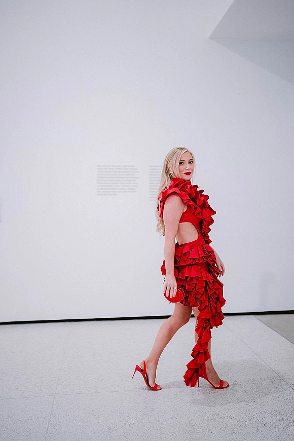 Wedding guest outfit in a red wedding guest dress with ruffle detail, matching heels and clutch, posed by a white gallery wall with text