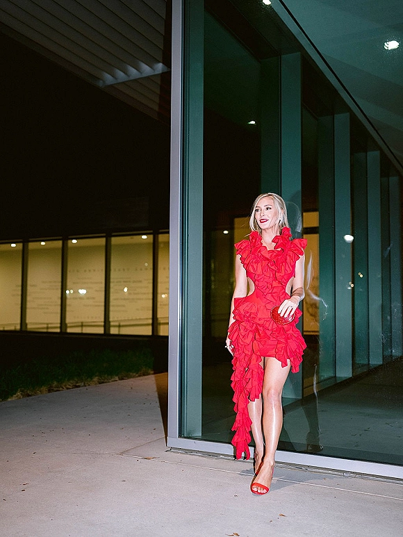Wedding guest outfit in a red wedding guest dress, holding a red clutch with matching heels by a glass-walled modern building at night