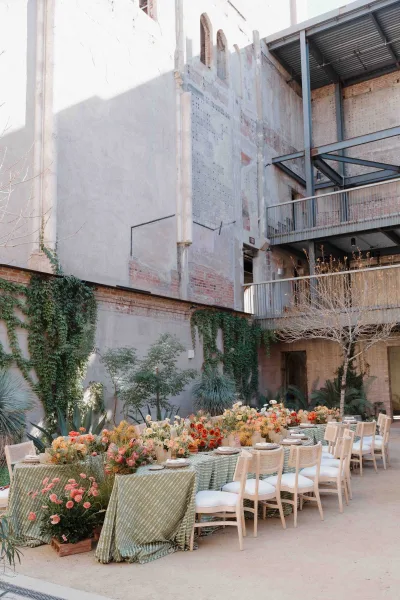 Reception tablescape with green patterned tablecloth, floral centerpieces and potted plants on a long banquet table in an ivy brick courtyard