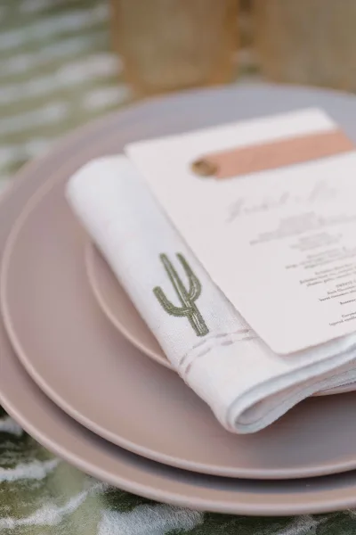 Wedding place setting with pink wedding plates, blush napkin, cactus embroidered napkin, menu card and leather place card in soft light