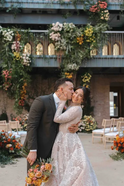 Wedding kiss portrait of bride and groom kiss beneath a floral arch, bouquet in hand, in a brick courtyard with arched windows