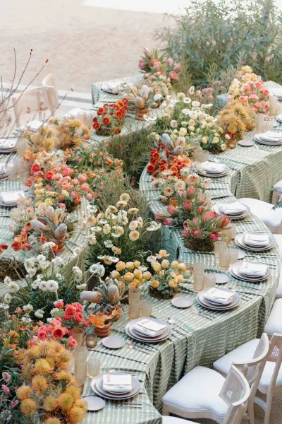 Reception tablescape with a green patterned tablecloth, wildflower centerpieces, and candlelit place settings on an outdoor patio with shrubs