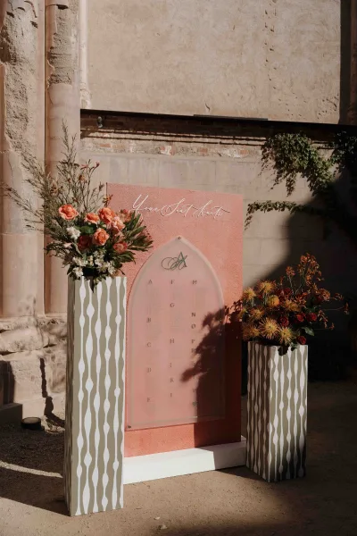 Wedding seating chart on an arched acrylic seating chart sign with coral backdrop and rose florals, set in a sunlit stone courtyard wall