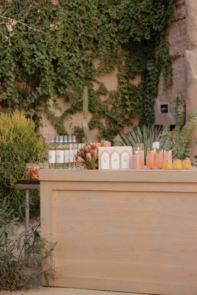 Wedding bar setup with outdoor wedding bar on a wood counter, pre-poured cocktails, wine bottles, and candlelit florals by an ivy wall and cactus backdrop