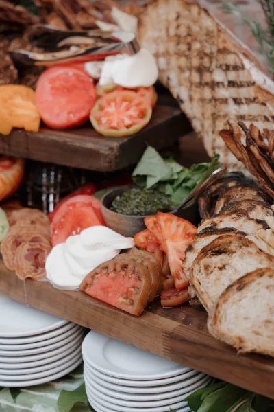 Wedding grazing table with rustic wood serving boards, bread, crackers, sliced tomatoes, herb dips, plates, and greenery on table surface