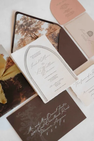 Invitation suite flatlay with deckle edge paper, calligraphy cards, RSVP, dark brown envelopes, vellum wrap and wax seal on white surface