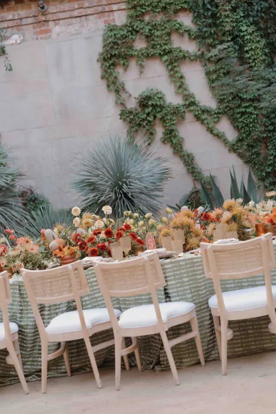 Reception tablescape on an outdoor reception table with rust florals, bud vases, taper candles, and place cards in an ivy stone courtyard.