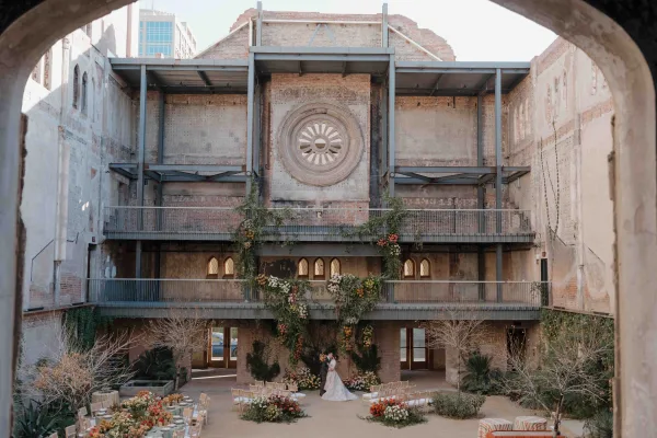 Wedding ceremony setup with an industrial wedding ceremony vibe, semicircle chairs facing a floral arch in a brick-walled courtyard with arched windows
