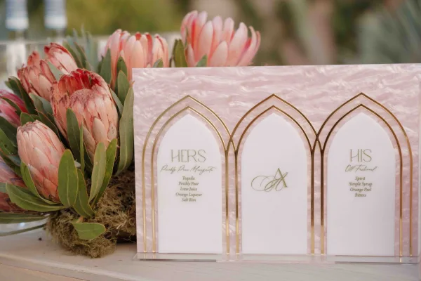 Signature drink sign with a his and hers drinks sign design in a gold frame on a wood tabletop, with protea flowers and greenery accents
