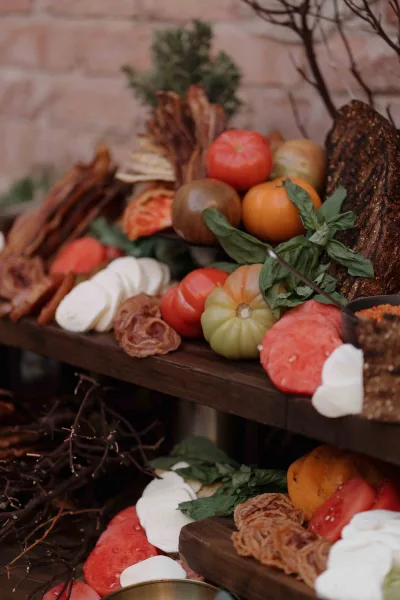 Wedding charcuterie board on a wooden serving board with tomatoes, basil, sliced bread, cheeses, and cured meat against a brick wall backdrop