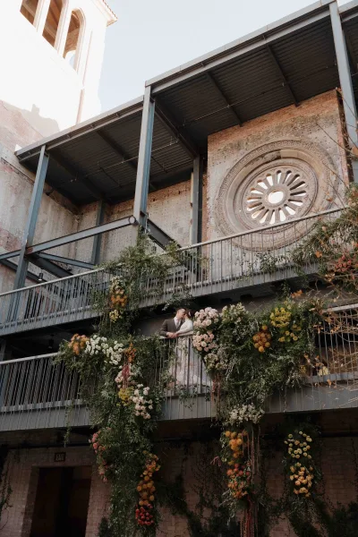 Wedding couple portrait of bride and groom kissing on a balcony railing with rose greenery garland, against industrial brick wall and round window