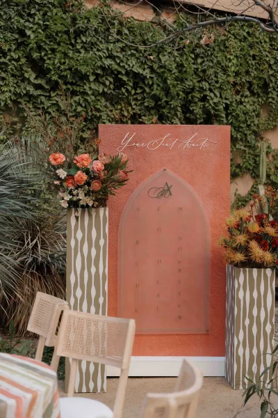 Wedding seating chart on an arched acrylic seating chart sign with calligraphy, flanked by coral desert florals and palm fronds on striped pedestals