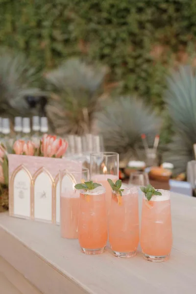 Wedding signature cocktails in highball glasses with ice and mint garnish, pink drinks beside menu cards and a candle on an outdoor bar counter