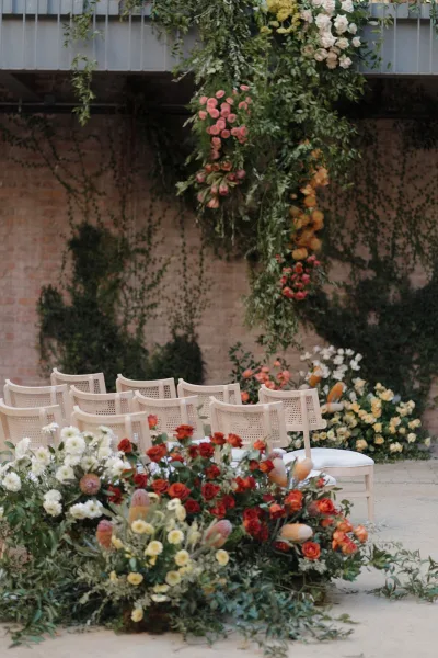 Ceremony setup with hanging floral installation over white chairs, framed by ground florals and greenery garlands in an ivy brick courtyard patio