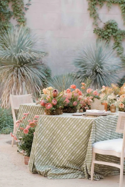 Reception tablescape with a wedding sweetheart table, green patterned tablecloth, low florals, moss runner, and votive candles by an ivy stone wall