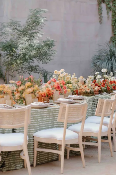 Reception tablescape with floral centerpieces and taper candles on a green patterned tablecloth, set on a courtyard patio by a stone wall