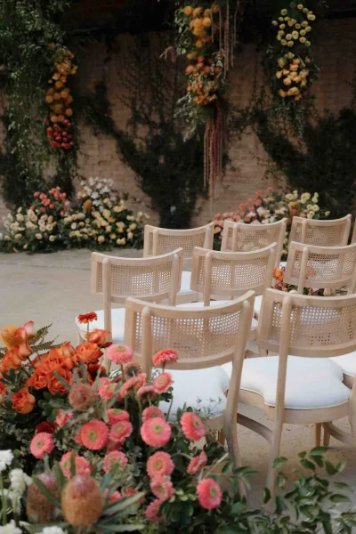 Ceremony setup with wedding ceremony seating of cane back wedding chairs, aisle florals, and hanging greenery against a brick wall backdrop
