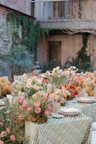 Reception tablescape with an outdoor reception table set on a long banquet, protea centerpieces and candles in glass votives in an ivy courtyard