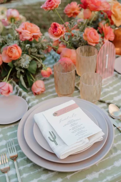 Reception tablescape with wedding place setting, coral rose centerpiece, amber glasses, striped tablecloth, and menu on napkin outdoors
