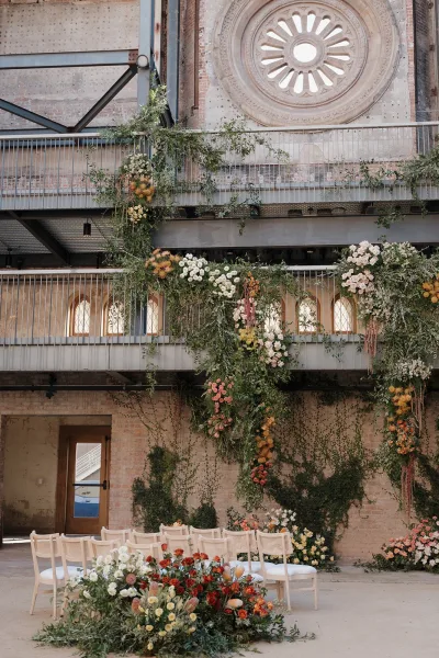 Ceremony floral installation with hanging wedding florals and trailing greenery garlands, set against an industrial brick wall and circular window backdrop
