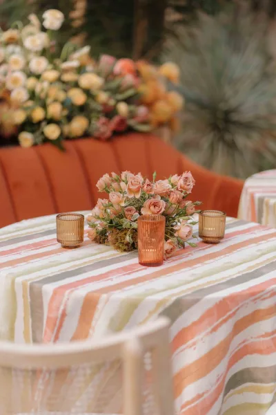 Reception tablescape with a striped wedding tablecloth, peach rose centerpiece, ribbed glass votives, and garden greenery with pampas grass backdrop