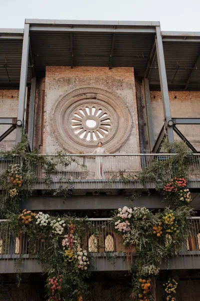 Bridal portrait of a bride on balcony in a lace long-sleeve gown, framed by greenery garlands and hanging florals on brick facade