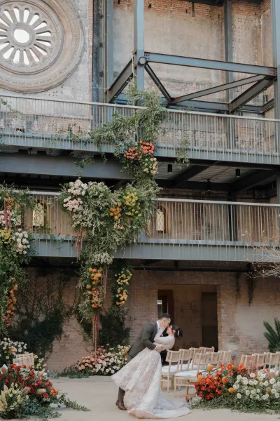 Wedding kiss portrait of bride and groom kiss beneath a floral arch with greenery garland, set against an industrial brick building and round window