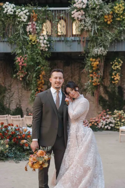 Couple portrait of bride leaning on groom, holding an orange-rose bouquet before a floral arch in a stone courtyard balcony setting