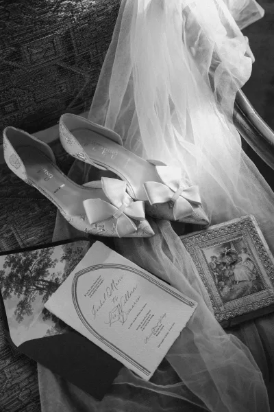 Wedding details flatlay with bridal shoes flatlay, bow heels, invitation suite, veil, and ring box styled on an upholstered chair