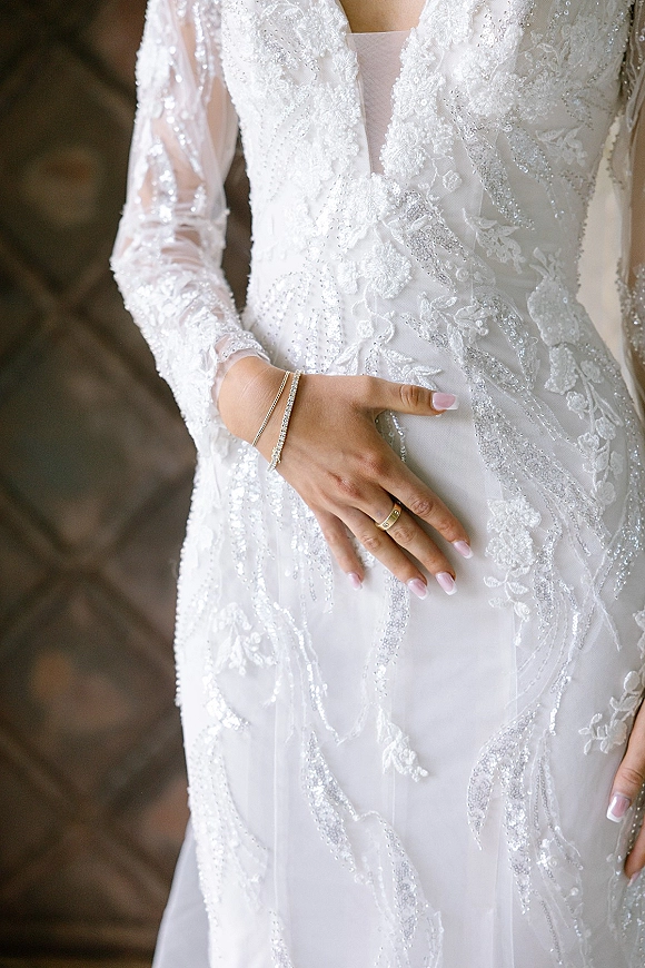 Wedding dress close-up showing beaded lace long sleeves and deep V neckline, bride’s ringed hand with bracelet against a stone wall