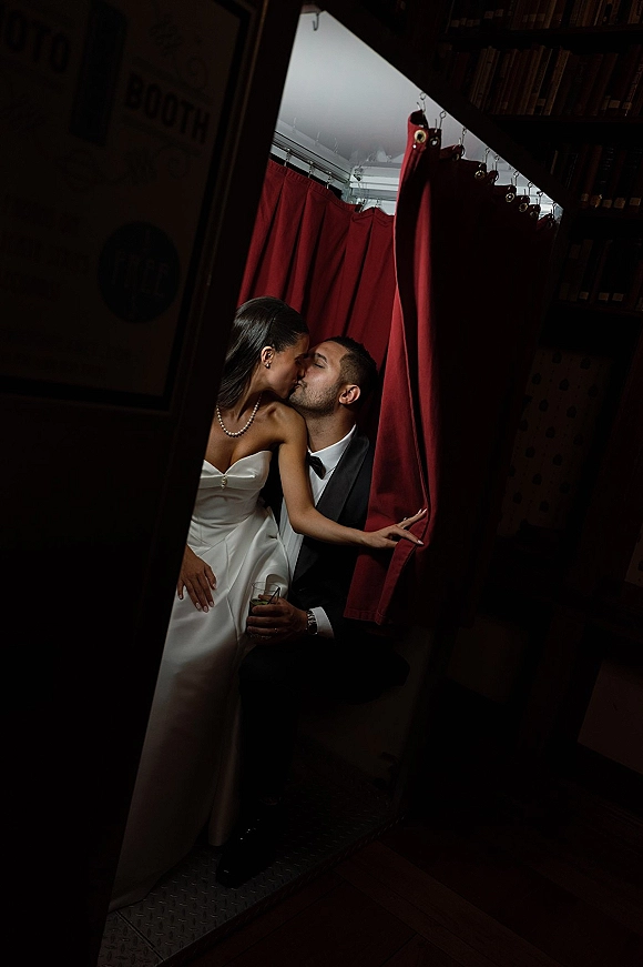 Wedding kiss portrait of bride and groom kissing in a narrow doorway, bride in pearl necklace and strapless dress, groom holding whiskey glass