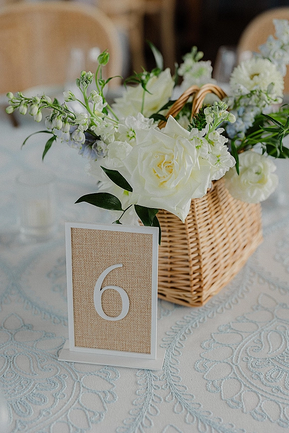 Reception centerpiece with a wicker basket of white roses and greenery beside table number 6 on a linen tablecloth, banquet chairs blurred behind