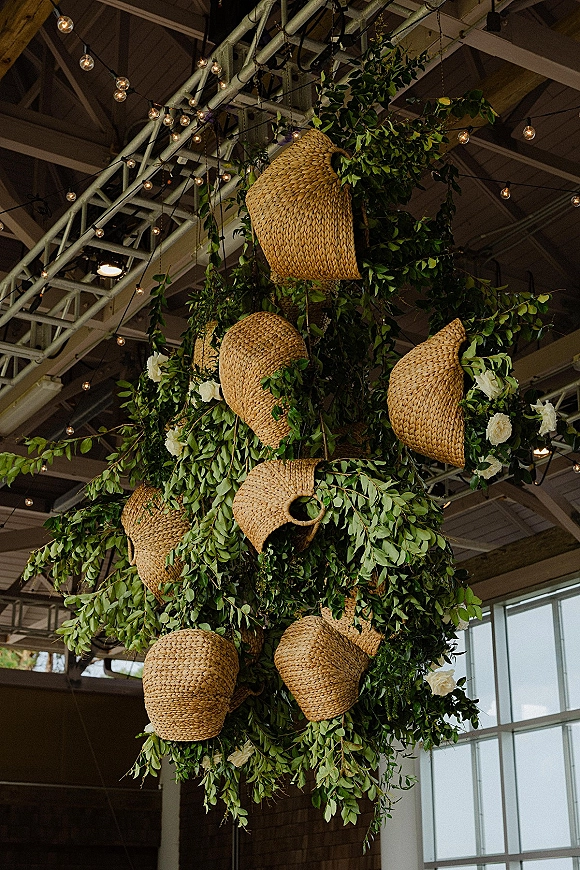 Hanging greenery installation with woven basket lantern accents, white roses and string lights suspended beneath industrial ceiling beams