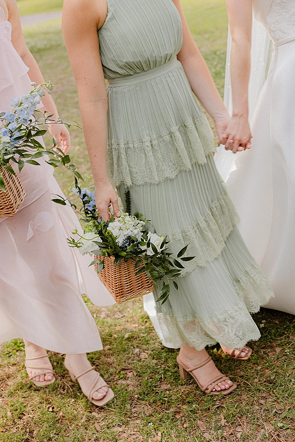 Bridesmaid dresses in sage green and blush pink with wicker flower baskets of blue and white blooms, lace trim and nude heels on grass lawn