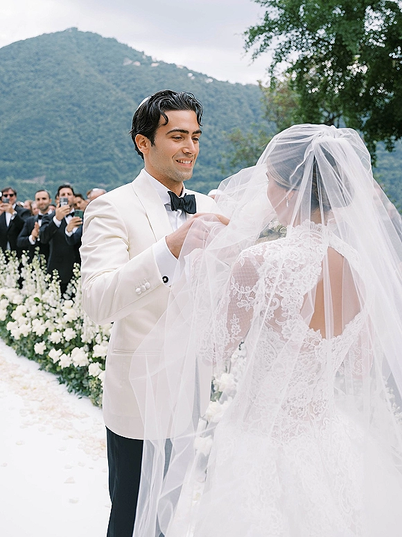 Wedding ceremony moment as groom lifting veil from bride in lace dress, white tuxedo and black bow tie amid aisle florals with mountain backdrop