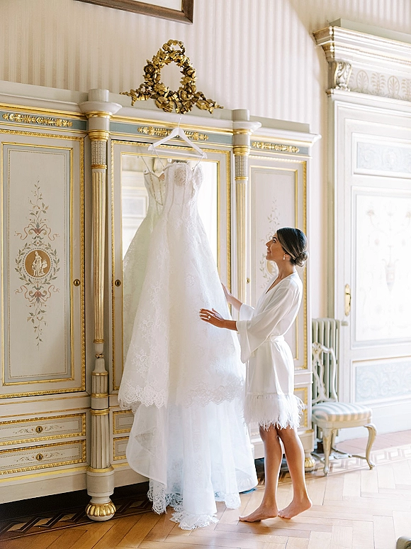 Wedding dress on a hanger, lace wedding dress details glowing in window light against an ornate wardrobe with gilded trim and wallpaper