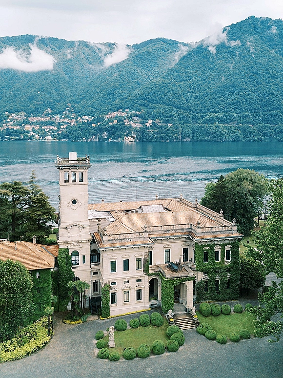 Wedding venue exterior with ivy-covered historic villa and bell tower, terracotta roofs, framed by manicured gardens and a mountain lake backdrop