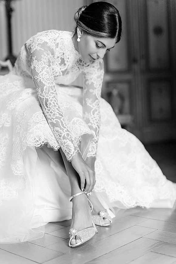 Bridal getting ready bride putting on shoes, adjusting glitter high heel sandals with an ankle bracelet on a wood floor by the door