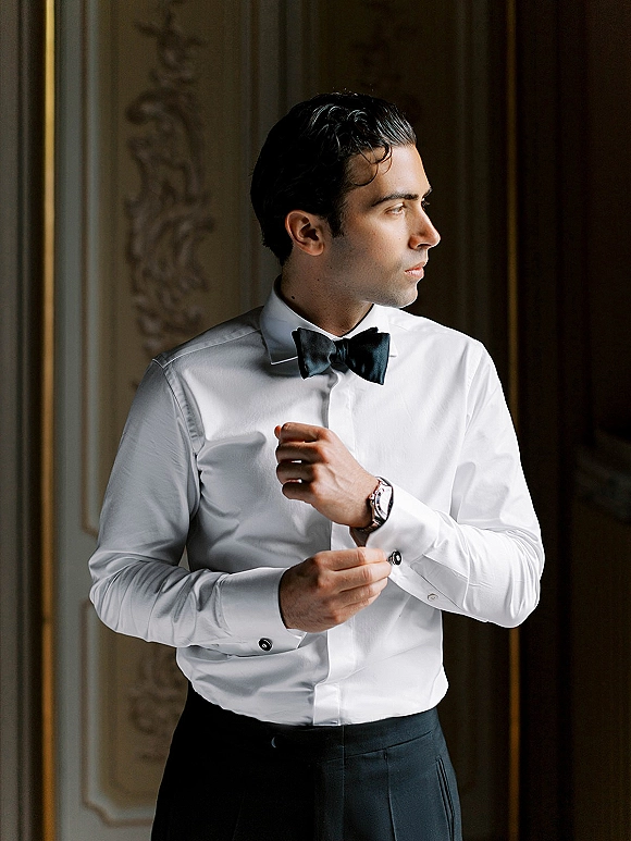 Groom portrait in a white dress shirt and black bow tie, adjusting cufflinks with a wristwatch by window light against ornate paneling