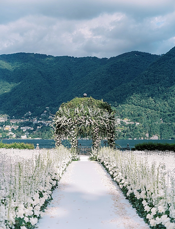 Ceremony aisle decor with a white aisle runner, flower-lined aisle and floral arch, set on a lakefront terrace with mountains beyond