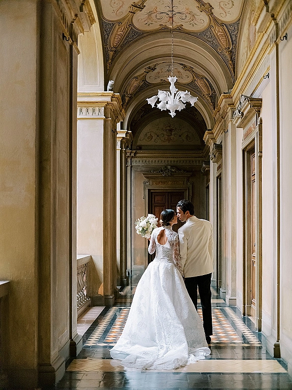 Couple portrait of bride and groom walking away, lace train and white bouquet in hand, beneath chandeliers in an ornate arched hallway