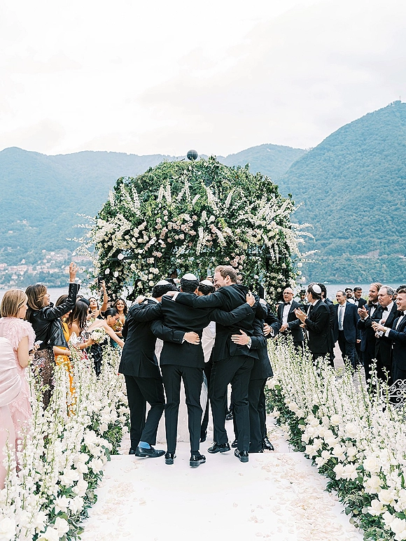 Ceremony moment at a Jewish wedding ceremony with guests cheering beneath a floral chuppah on a lakeside terrace with mountains beyond