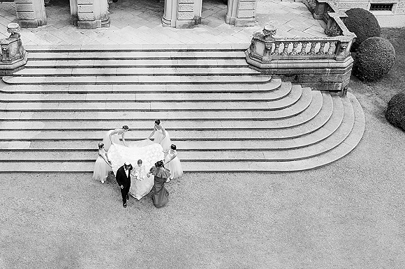 Bride entrance as bridesmaids adjust her long veil and train on a stone staircase, bouquet in hand, groom in tuxedo below.