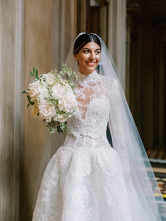 Bridal portrait of a bride holding bouquet of white roses and greenery, wearing a cathedral veil in a stone-walled indoor hallway