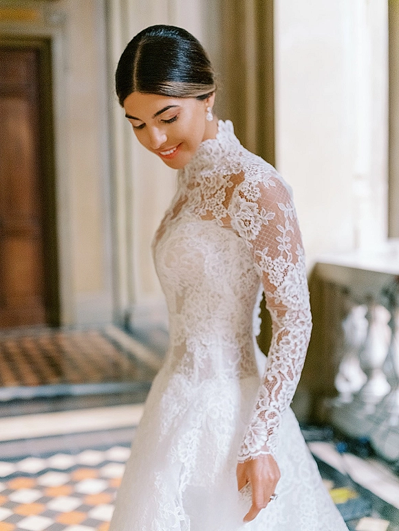 Bridal portrait in a lace wedding dress, bride looking down in an indoor hallway with window light, patterned tile floor and stone balustrade